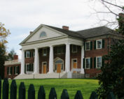 A large brick mansion with white columns behind a green fence.