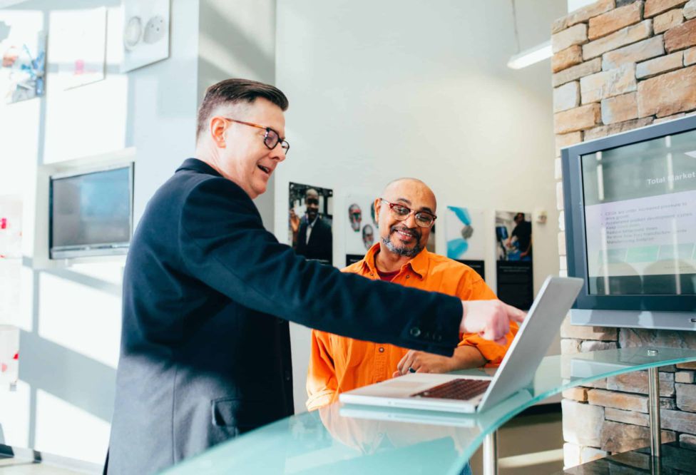 A man in a blazer points at a computer screen while a man in a polo shirt looks on and smiles.